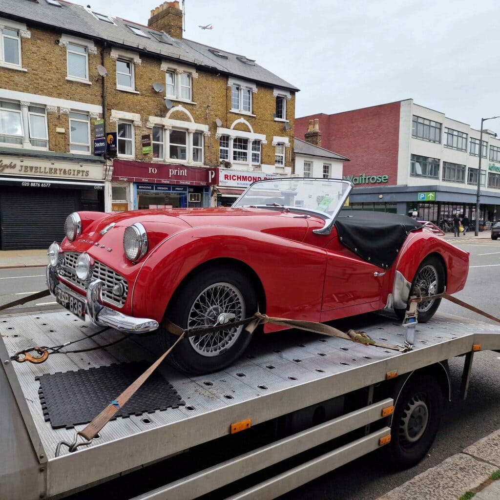A classic red Triumph TR3 convertible securely loaded onto a flatbed recovery truck on Richmond High Street, showing local breakdown recovery.
