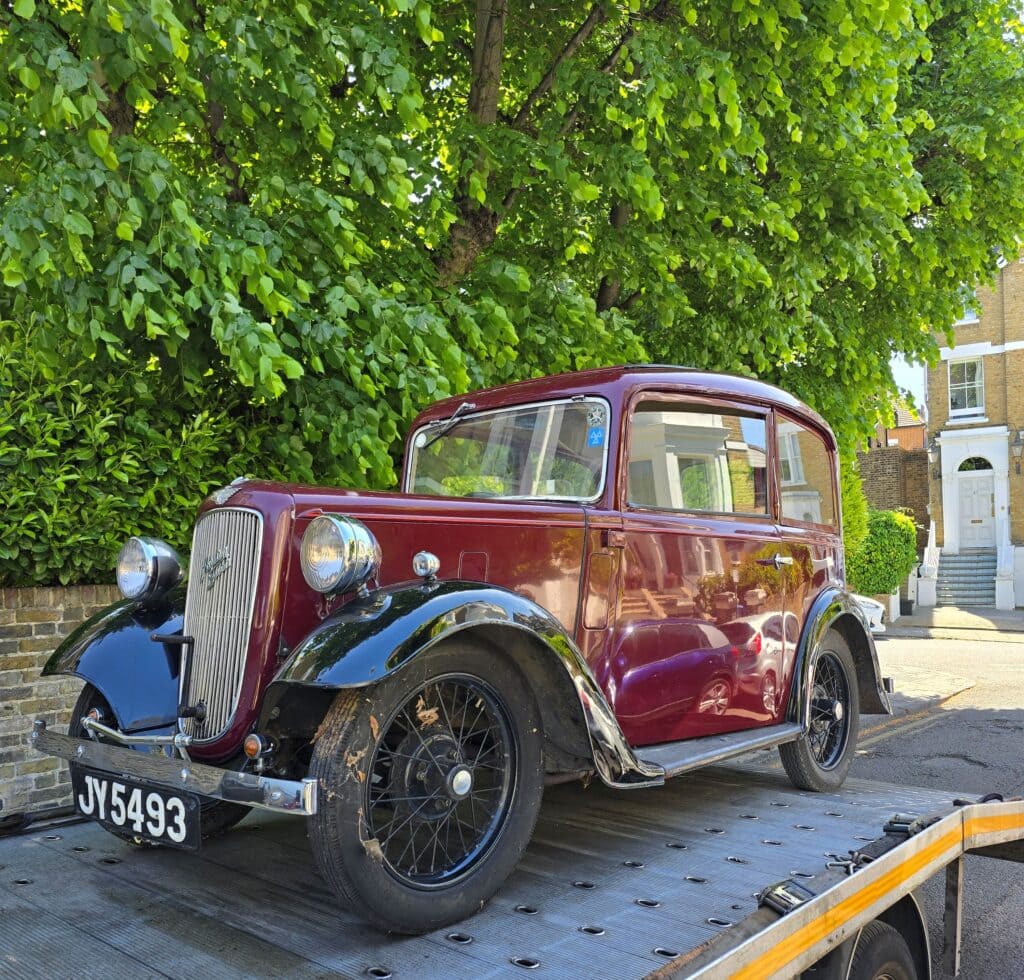 A maroon vintage Austin saloon securely loaded onto a flatbed recovery truck on a sunny residential street, demonstrating careful vehicle transport.