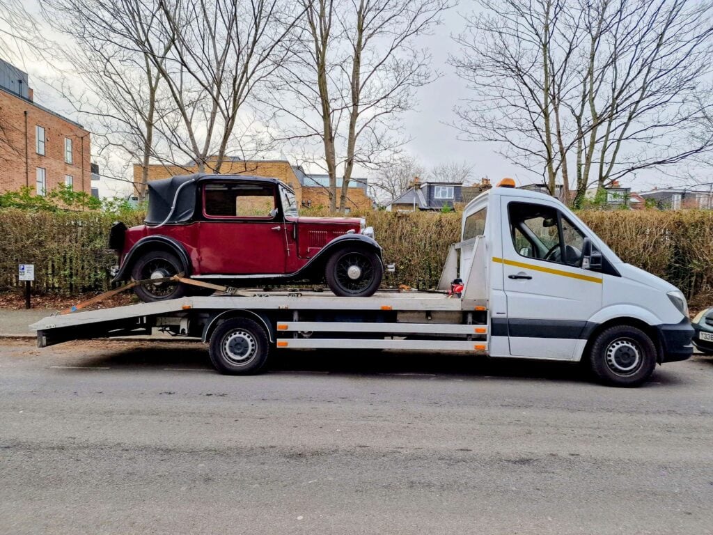 Side profile of a maroon vintage Austin Seven Ruby securely loaded on a flatbed truck, showcasing specialist transport for historic vehicles.