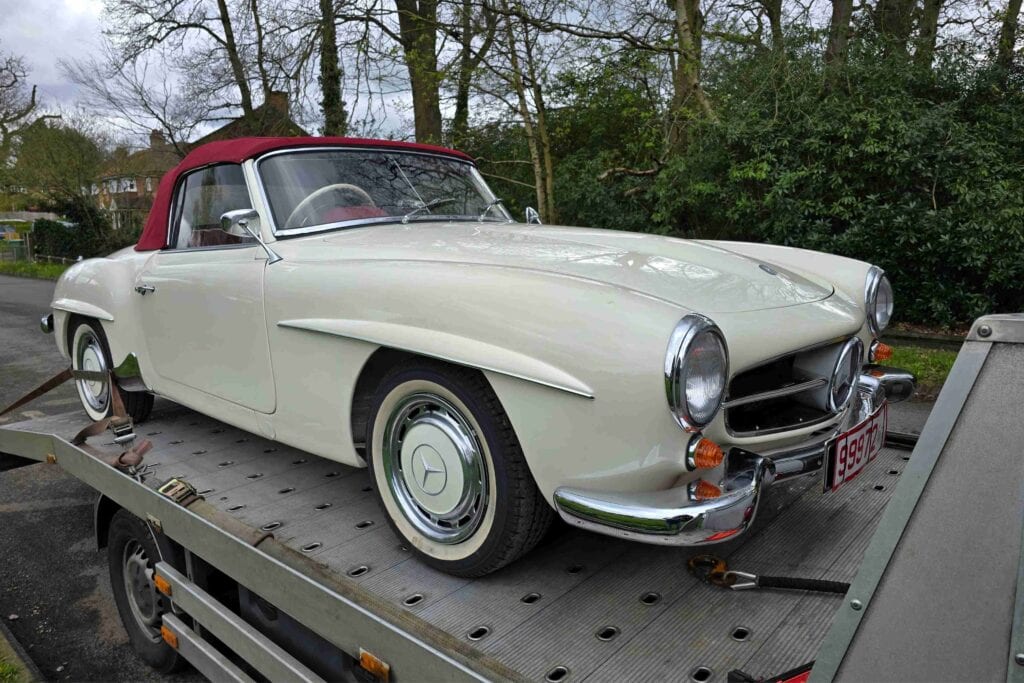 A classic white Mercedes-Benz 190SL with a red roof securely loaded onto a flatbed recovery truck on a quiet road.