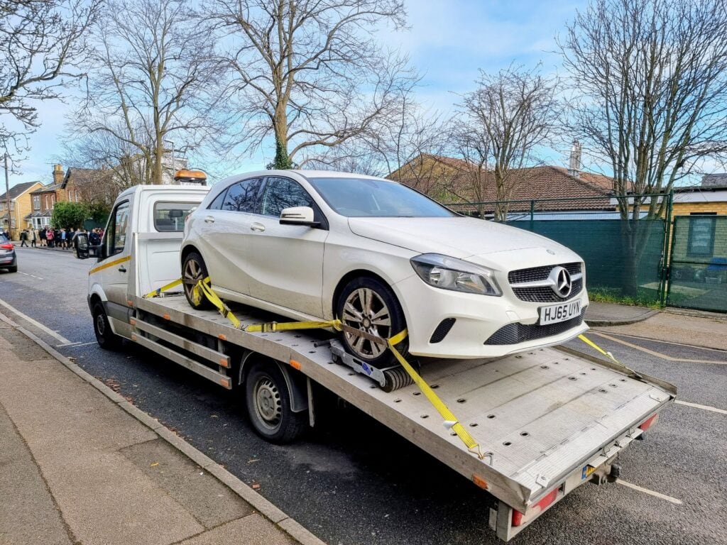A white Mercedes-Benz A-Class being loaded onto a flatbed recovery truck using wheel skates on a London street.