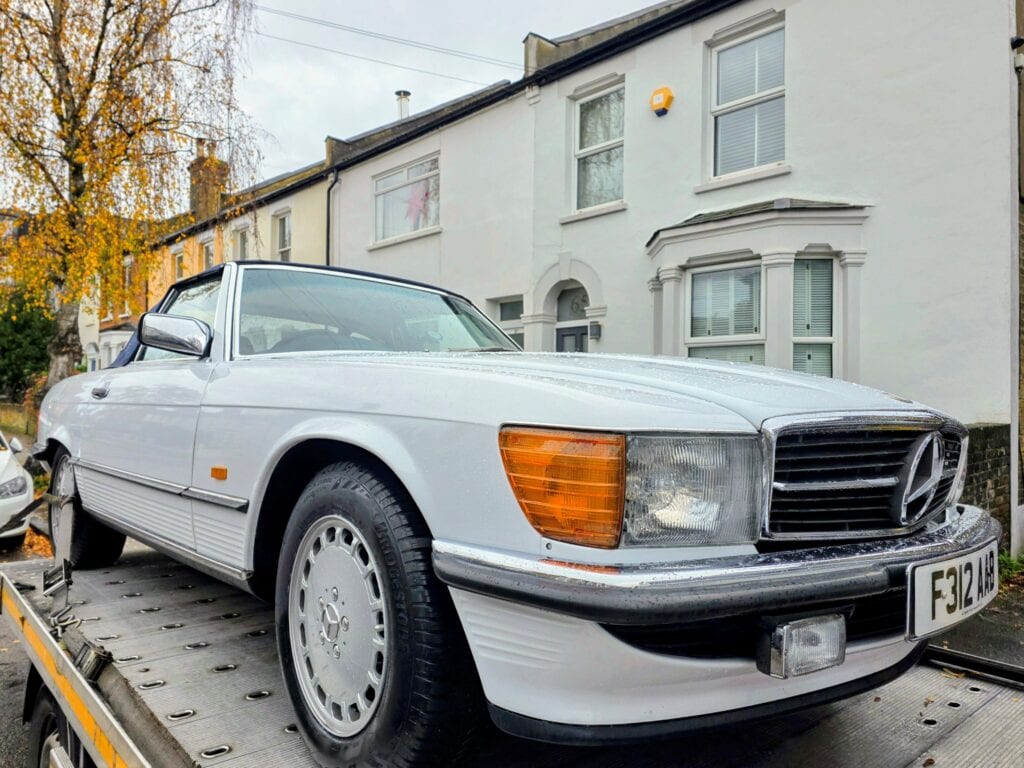 A classic white Mercedes-Benz R107 SL convertible restoration project on a flatbed recovery truck in a residential London street.