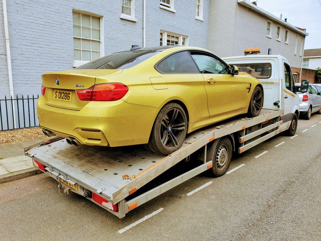 A yellow BMW M4 coupe secured on a flatbed recovery truck on a residential street in London.
