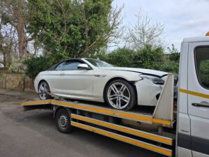 Damaged white BMW 6 Series being recovered on a flatbed tow truck in Twickenham after a roadside accident.