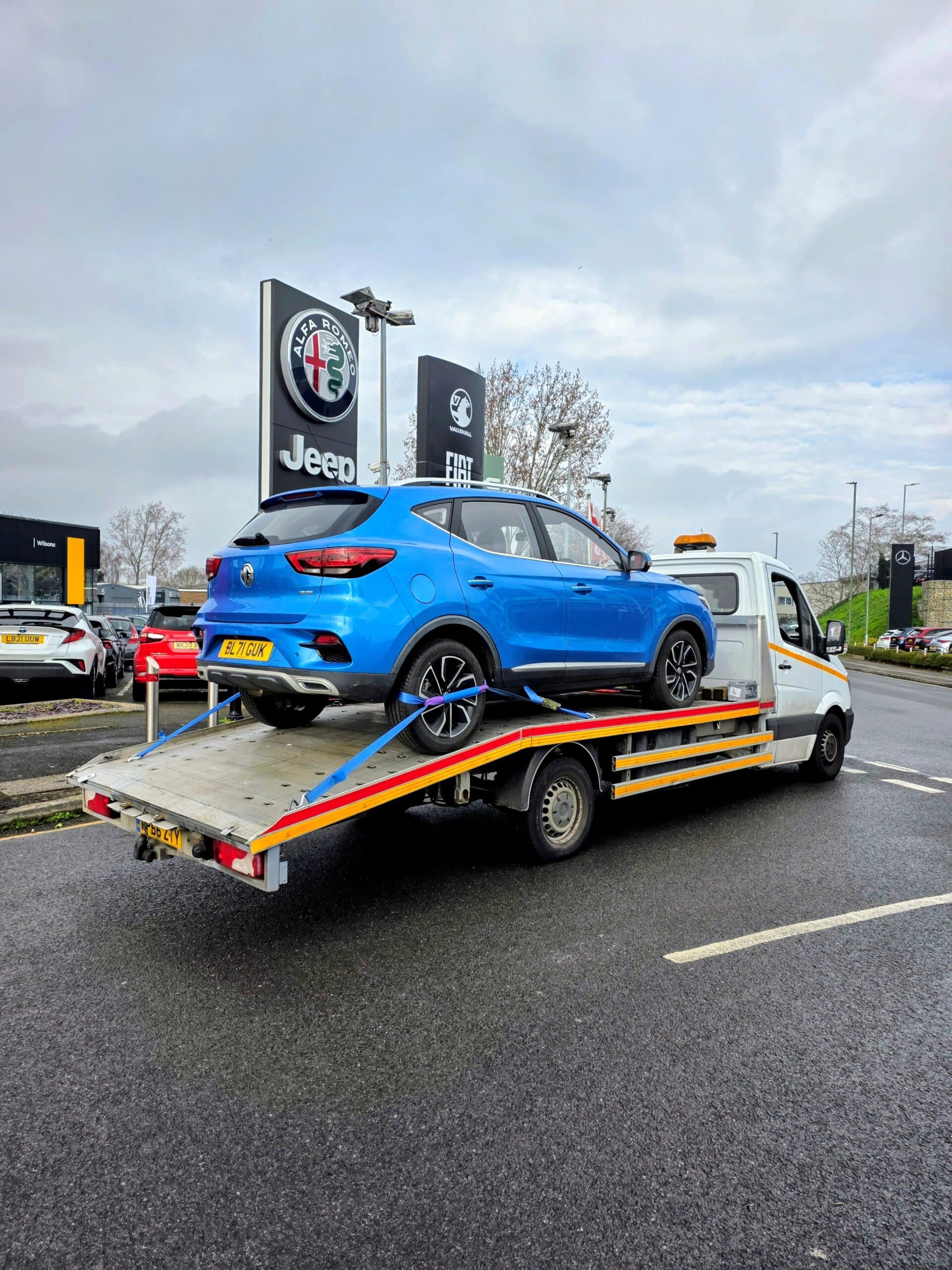 Professional car recovery Twickenham showing a blue MG ZS safely secured with blue wheel straps on a flatbed tow truck near a local dealership.