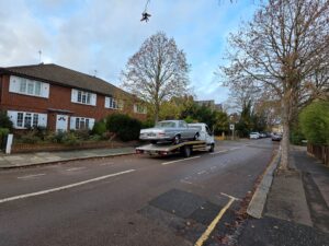 Professional long distance transport of a classic silver Mercedes-Benz Coupe on a flatbed recovery truck through a residential area.
