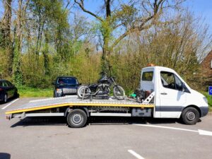 Professional motorbike recovery Twickenham featuring a black custom bobber motorcycle secured on a white flatbed recovery truck in a wooded parking area.