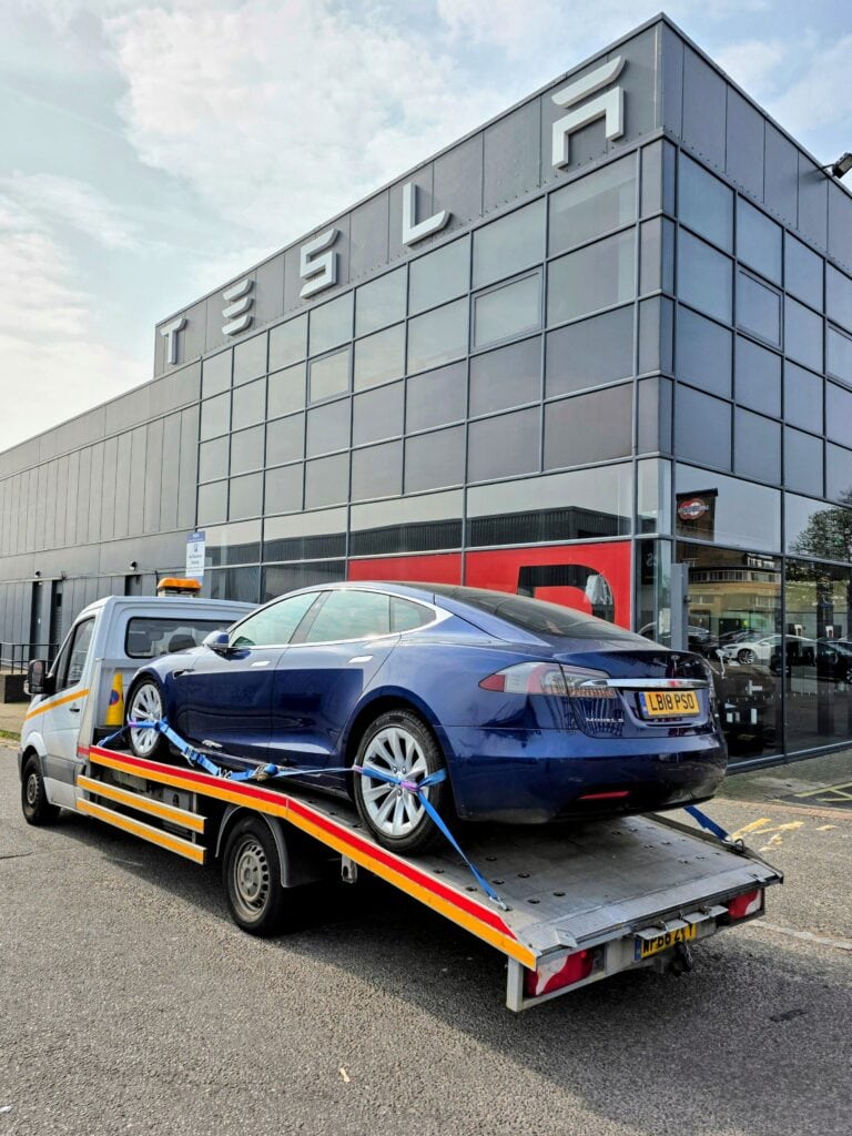 Professional car recovery showing a blue Tesla Model S secured on a flatbed tow truck parked outside a modern Tesla service building.