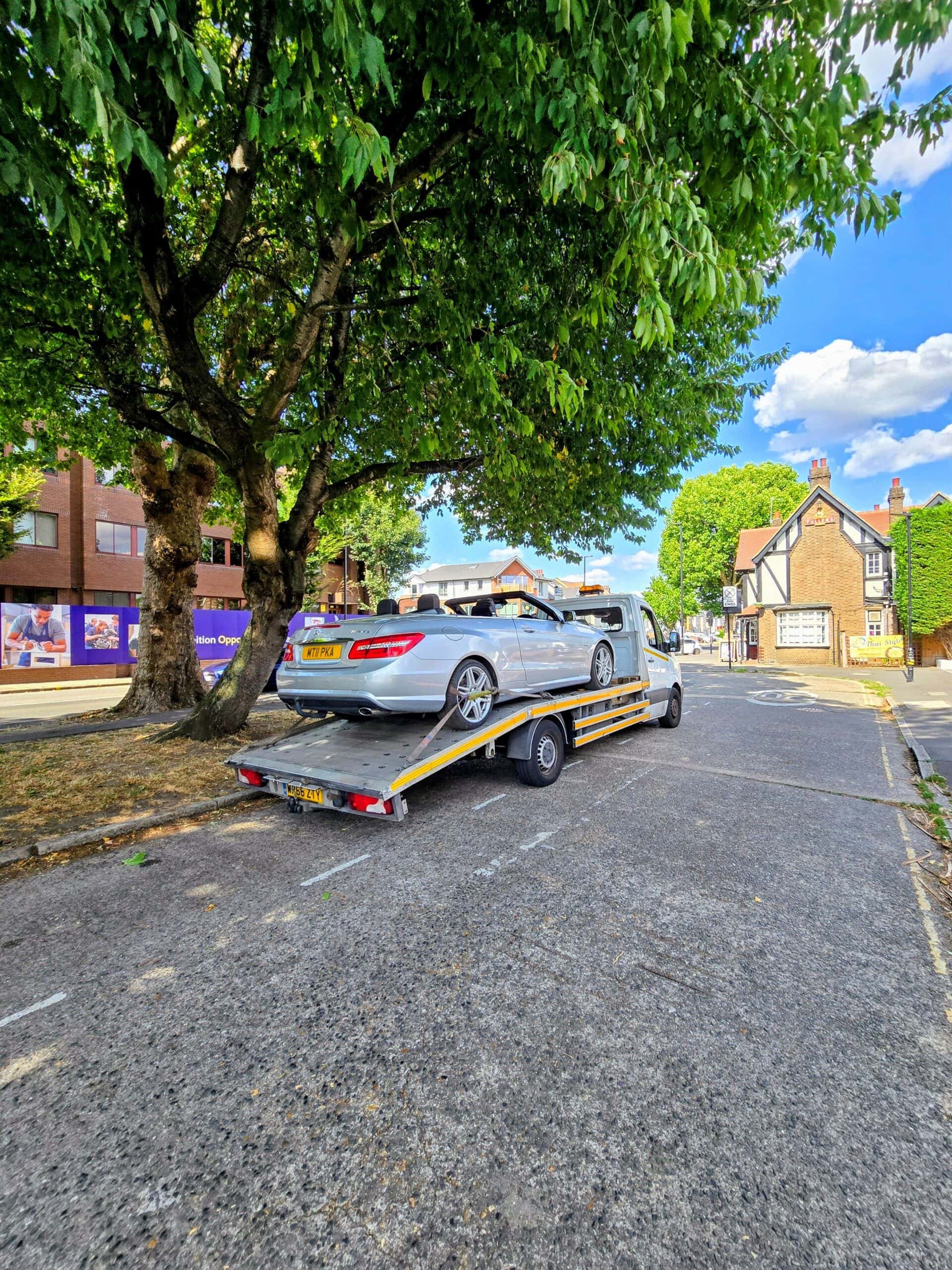White flatbed tow truck with yellow chevrons carrying a silver Mercedes E-Class Cabriolet on a London street with trees.