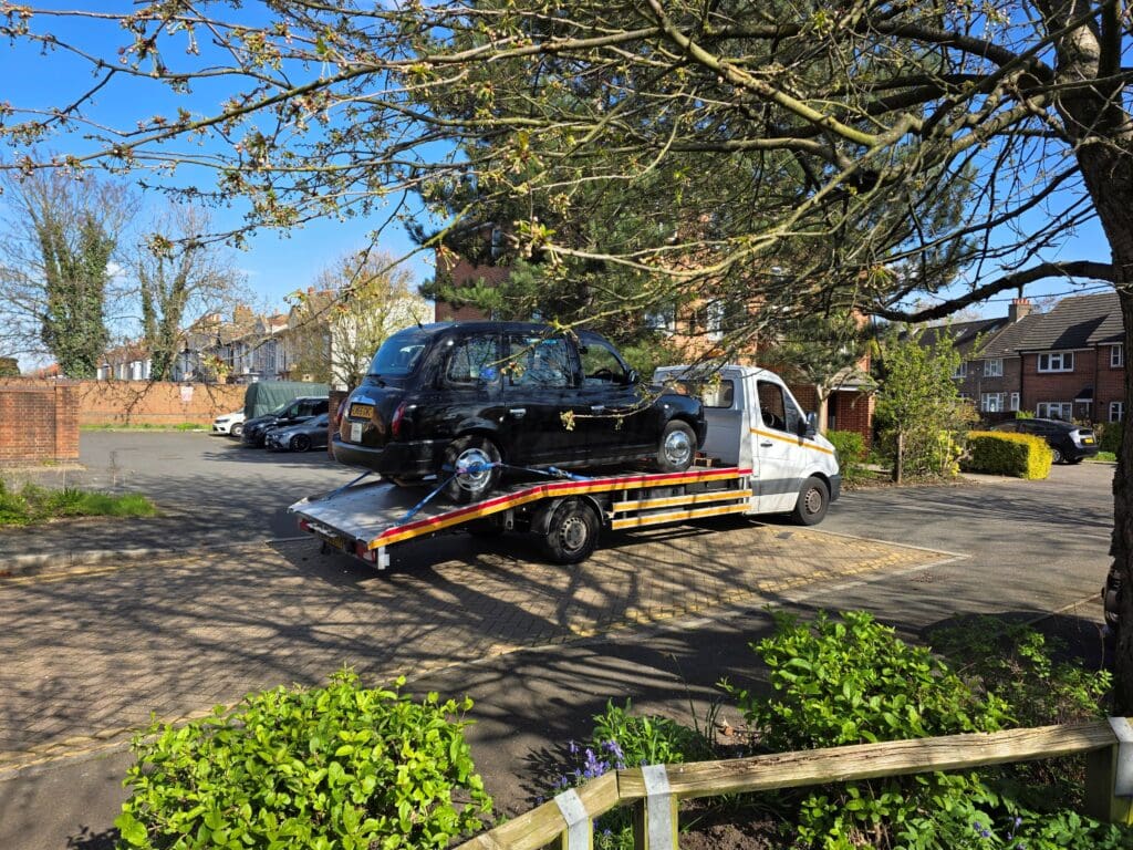 A black LTI TX4 London Taxi secured on a flatbed recovery truck for professional breakdown recovery and towing service in Wimbledon SW19 area.