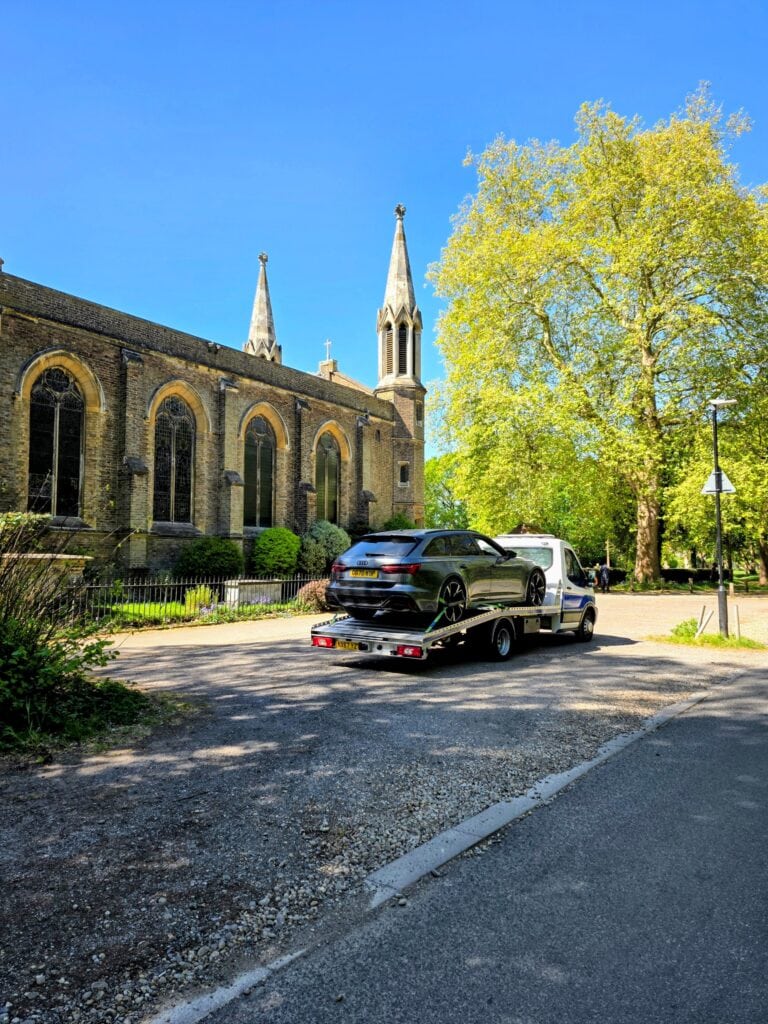 A grey Audi RS6 being transported on a London Auto Towing flatbed truck past a historic Southwest London church.