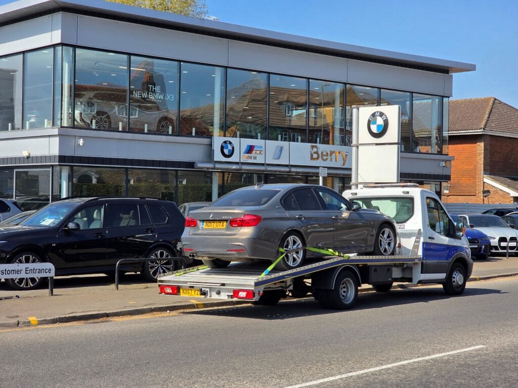 A grey BMW 3 Series sedan securely loaded on a flatbed recovery truck outside a local dealership during a professional towing service in Chiswick W4.